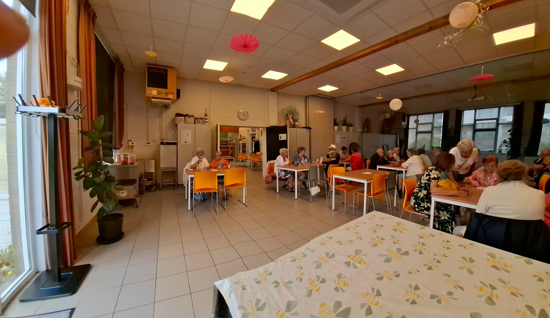 Grote gemeenschapsruimte met lange tafels, oranje stoelen en senioren die samen eten, met plafondlampen.